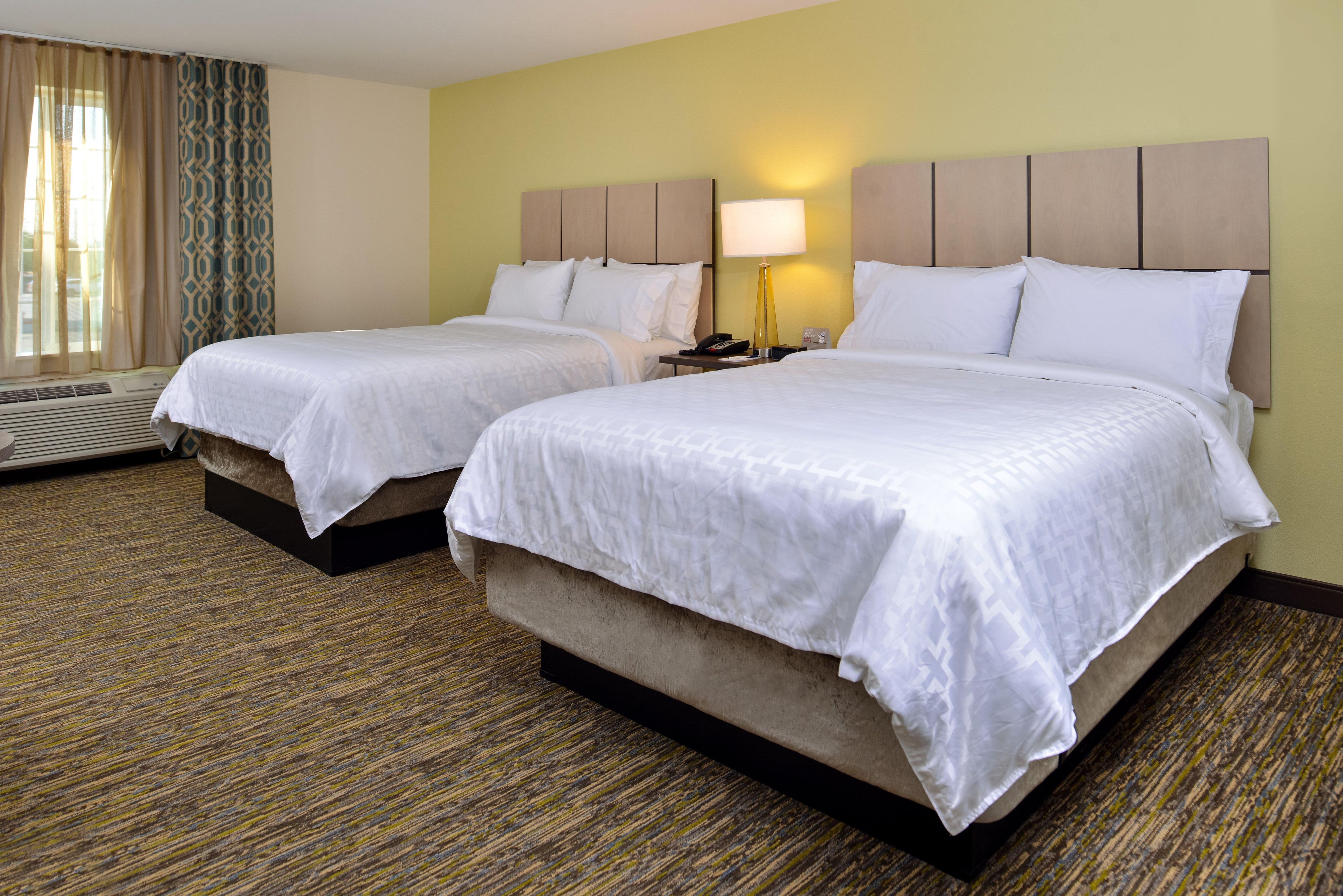 A hotel room with two beds, a bedside table with a lamp, and a window with curtains.