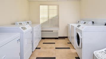 Une buanderie équipée de quatre machines à laver et d’un sèche-linge, avec un sol carrelé et une fenêtre dotée de stores.