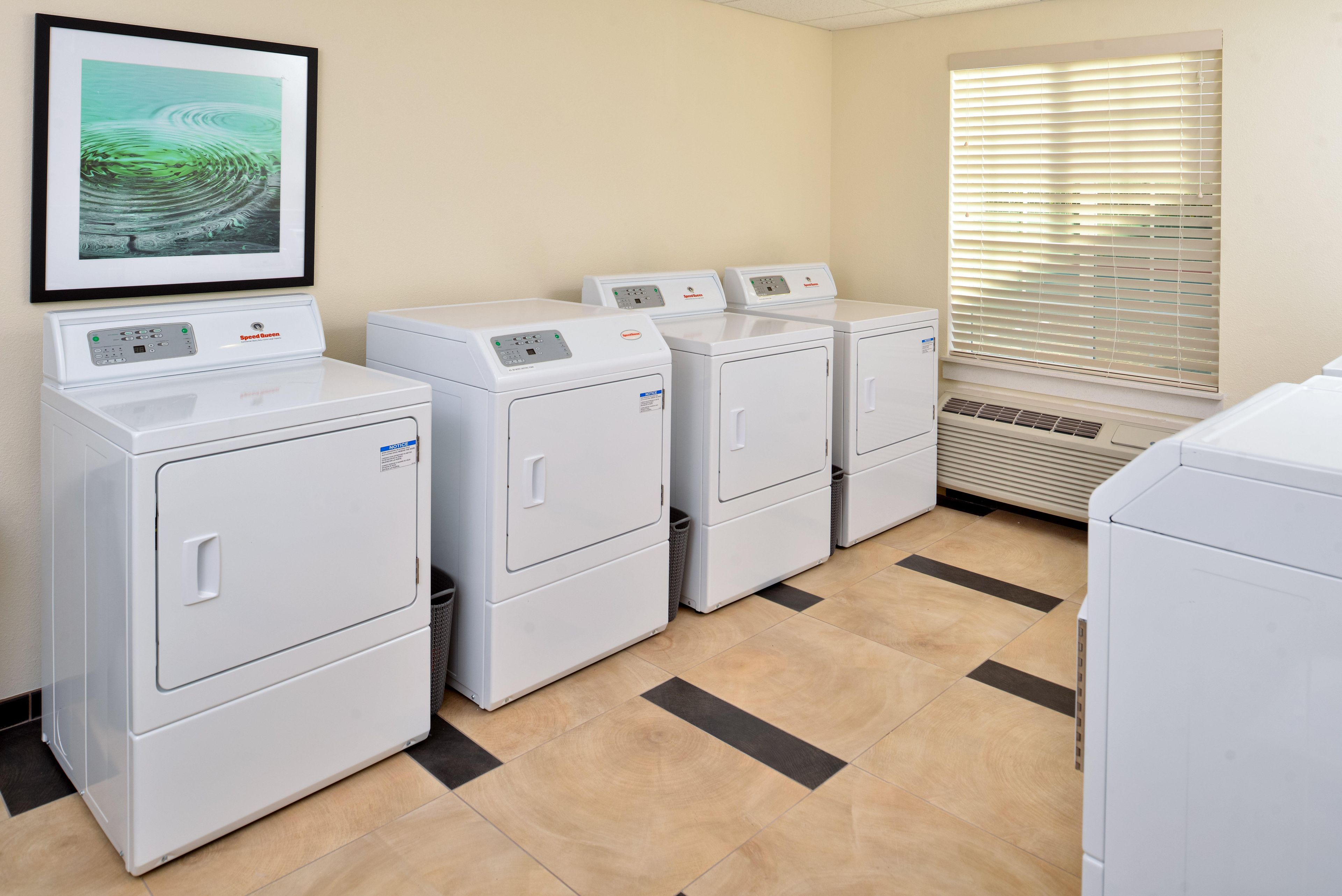 A laundry room with four stacked washers and dryers, a framed picture on the wall, and a window with blinds.