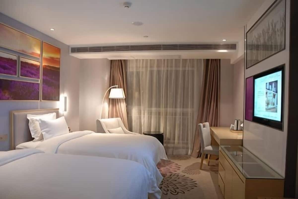 Ein Hotelzimmer mit zwei Betten, einem Schreibtisch, einem Stuhl, einem Fernseher und einem großen Fenster mit Vorhängen.