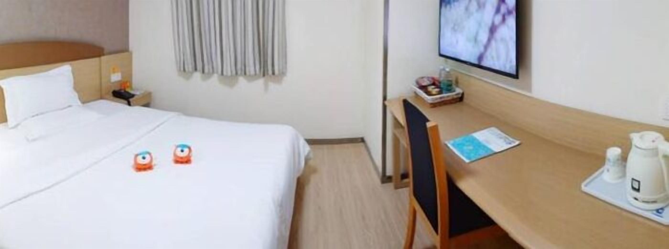 A hotel room with a bed, a desk with a chair, a television, and a window with curtains.