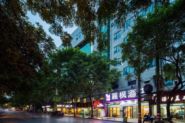 A street view at dusk with a building featuring a neon sign and a restaurant with outdoor seating.