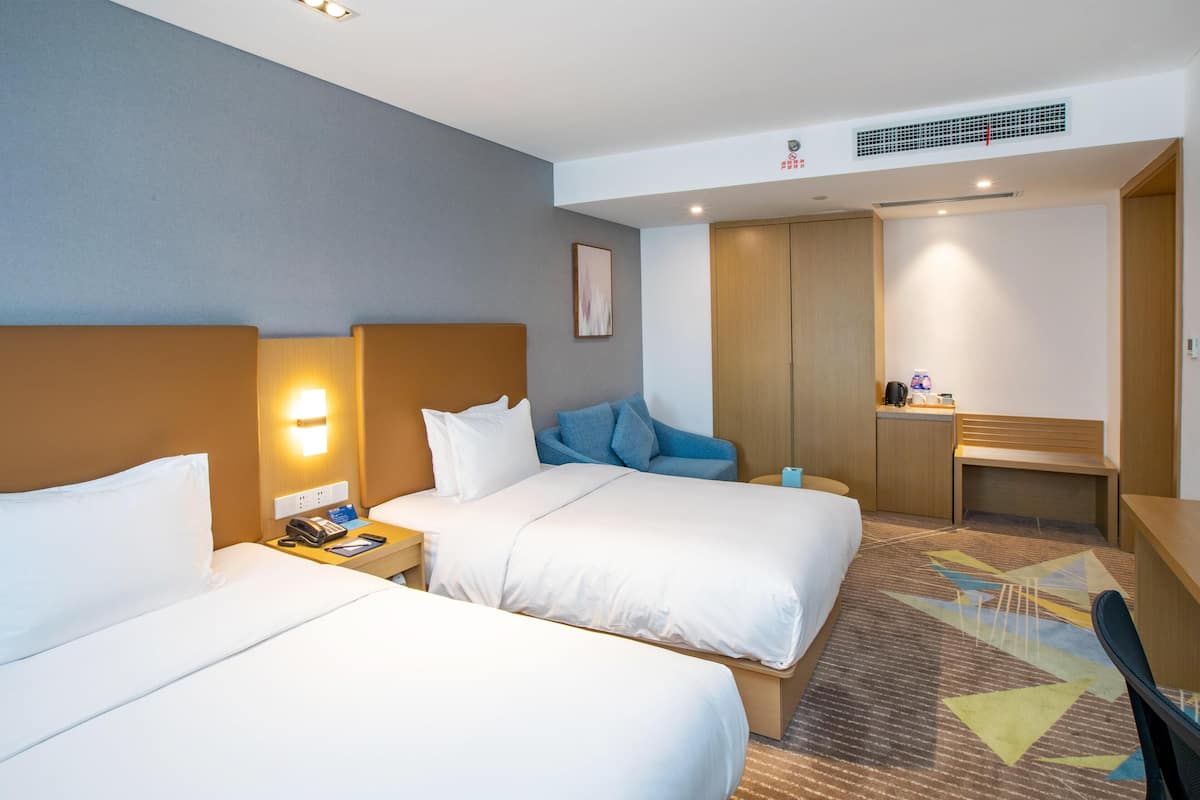 A hotel room with two beds, a desk, a chair, a nightstand with a phone, and a wall-mounted light.