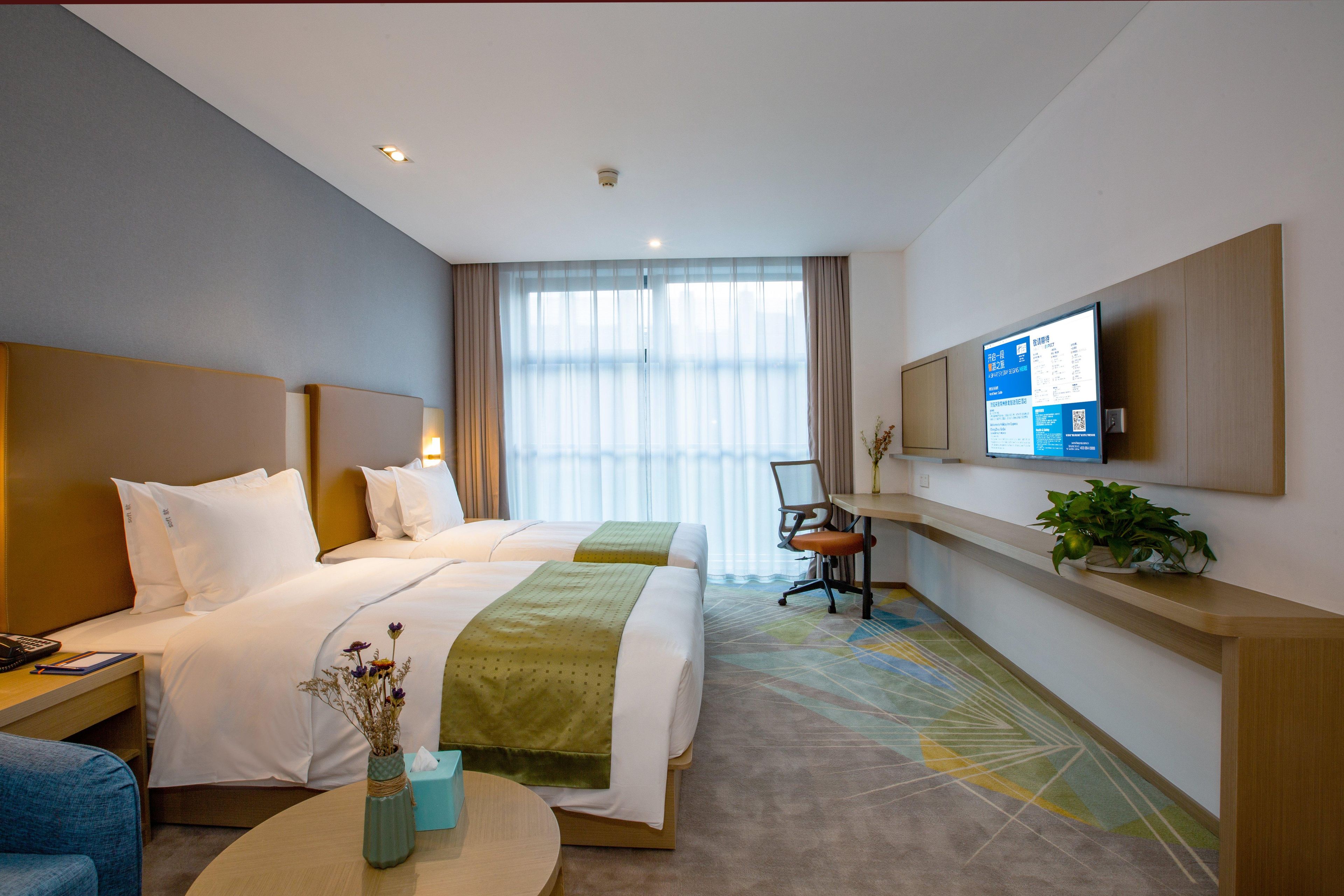 A hotel room with two beds, a desk, a chair, a TV, and a plant.