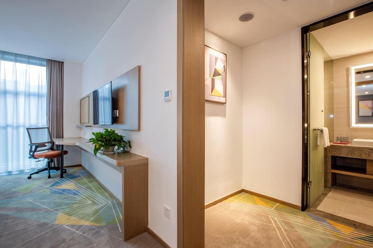 A hotel room with a desk, chair, potted plant, and a large window with curtains.