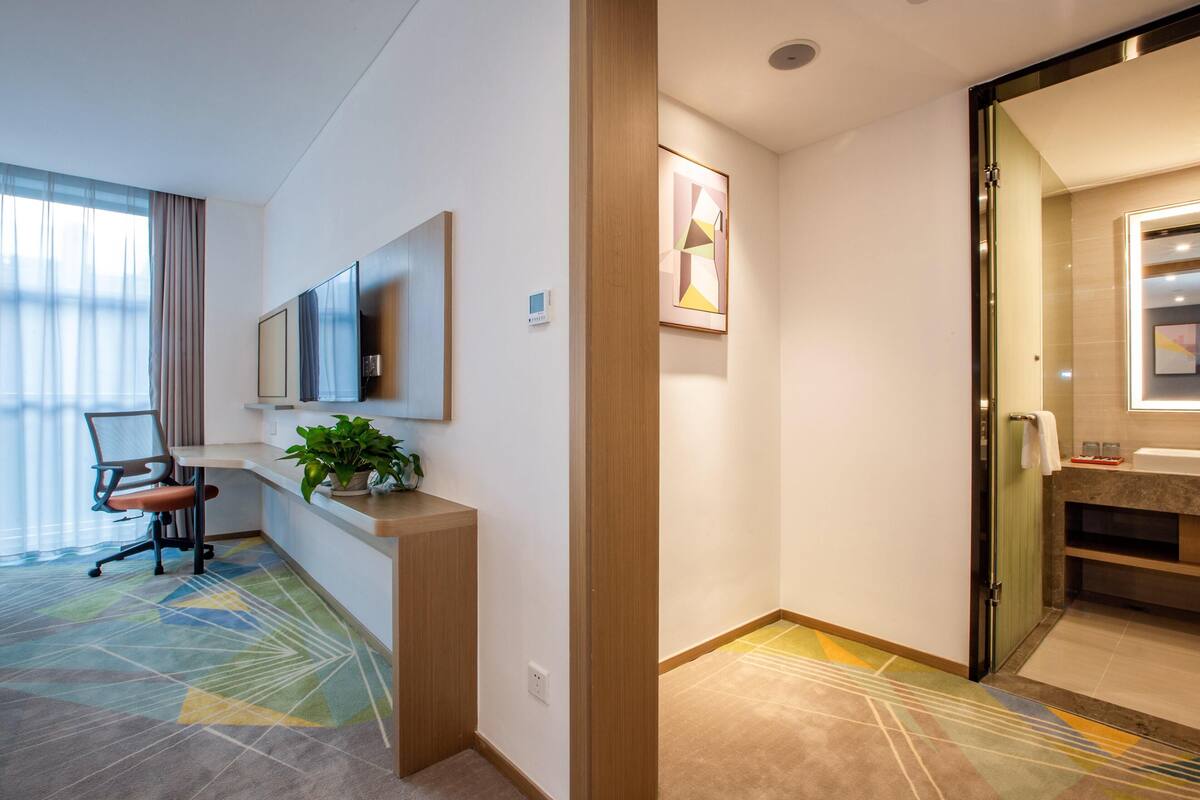 A hotel room with a desk, chair, potted plant, and a large window with curtains.
