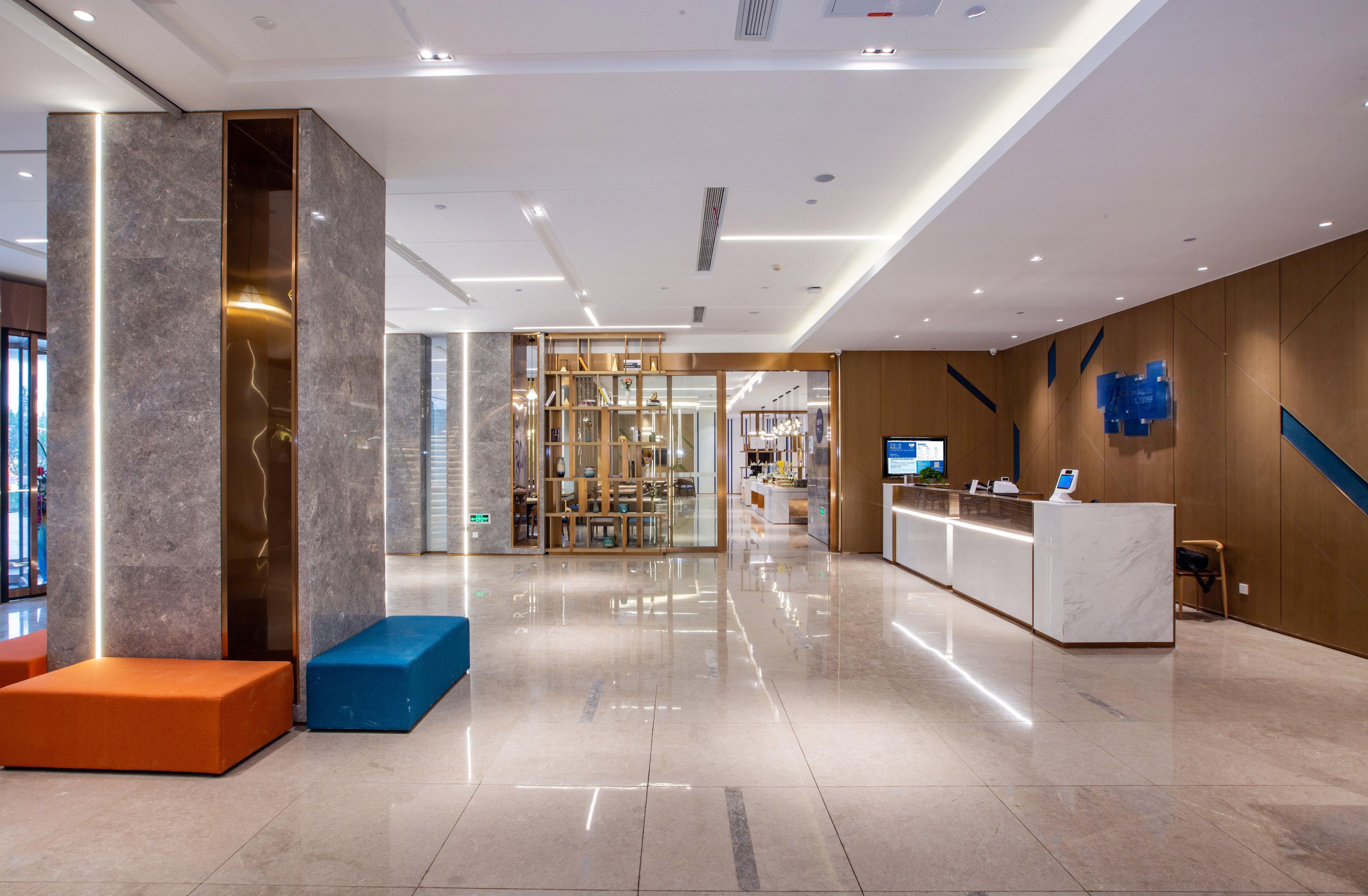 Un moderno vestíbulo de hotel con una recepción, zonas de estar y un separador de vidrio que conduce a un restaurante.