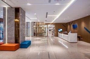 Un moderno vestíbulo de hotel con una recepción, zonas de estar y un separador de vidrio que conduce a un restaurante.