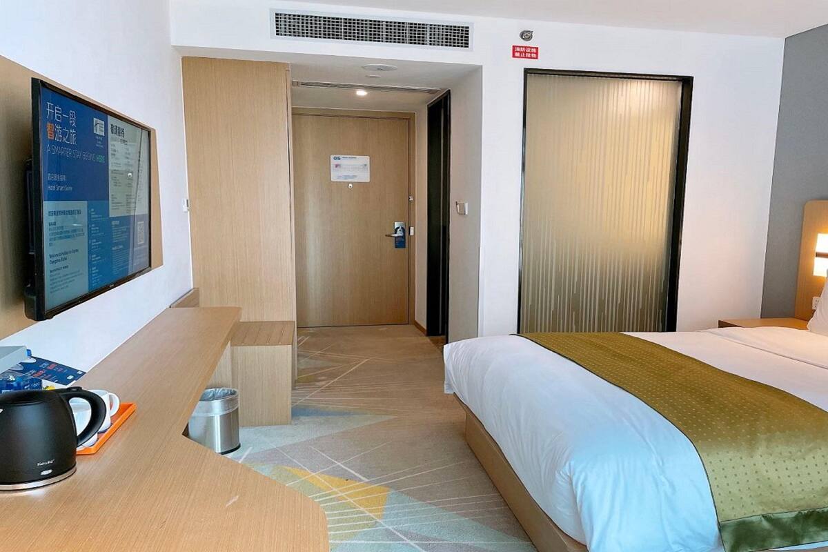 A hotel room with a bed, a desk with a coffee maker, a TV, and a door leading to another room.