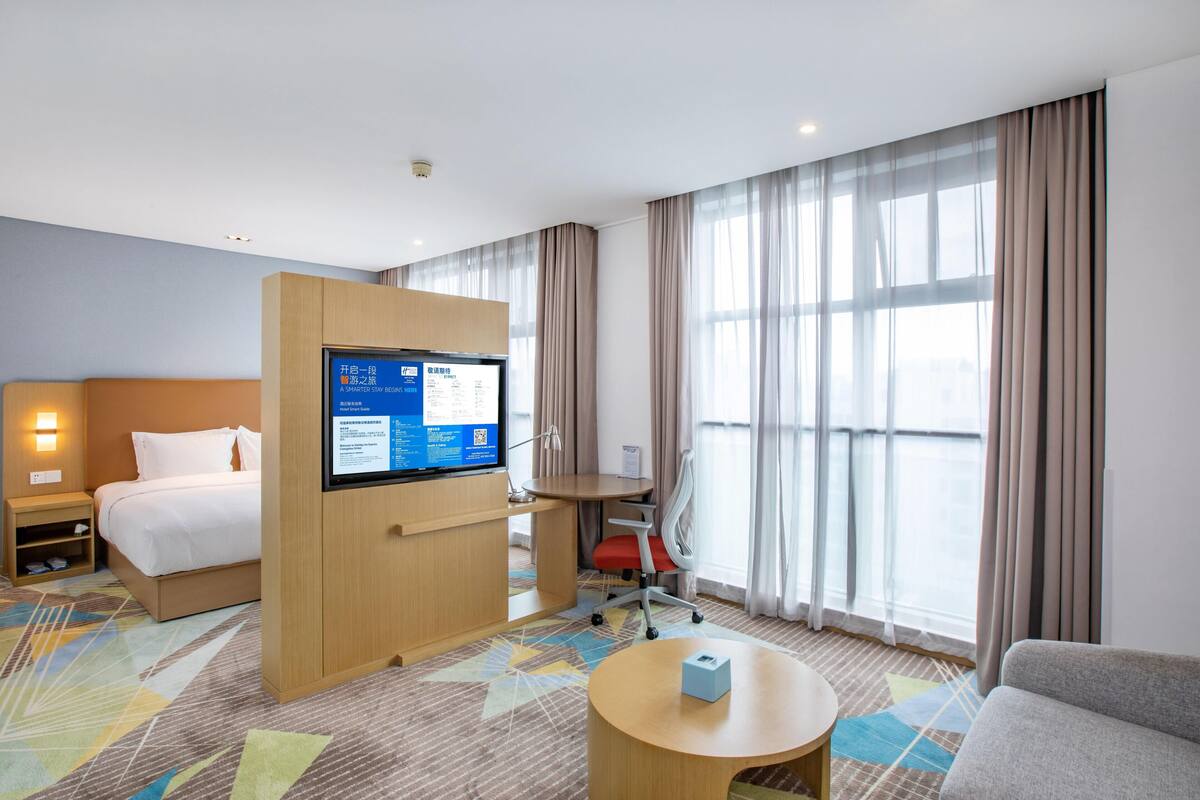 A hotel room with a bed, a desk with a computer, a chair, a small table, and a large window with curtains.
