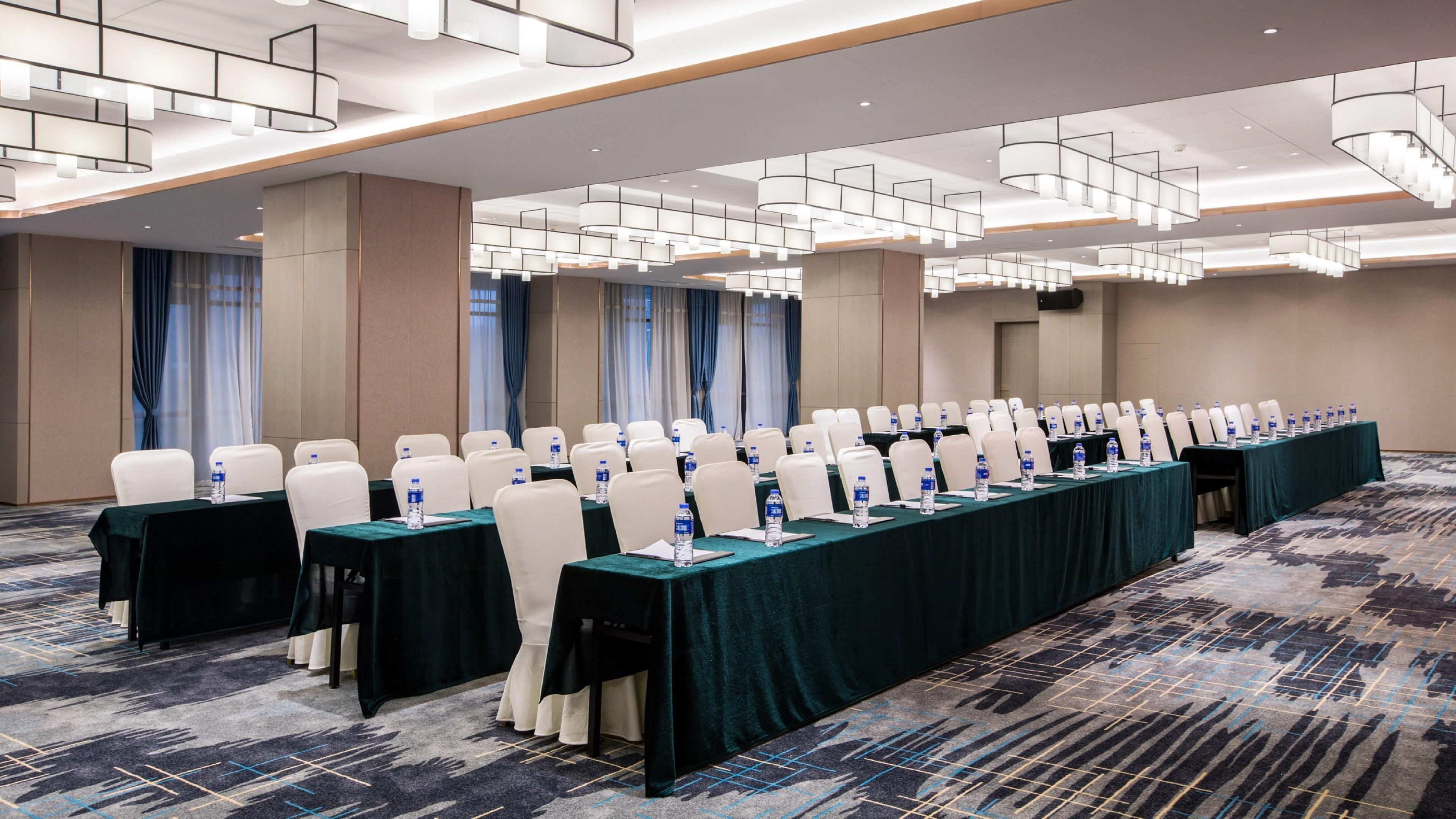 Un salón de conferencias con filas de mesas, sillas y botellas de agua, con una alfombra estampada e iluminación moderna.