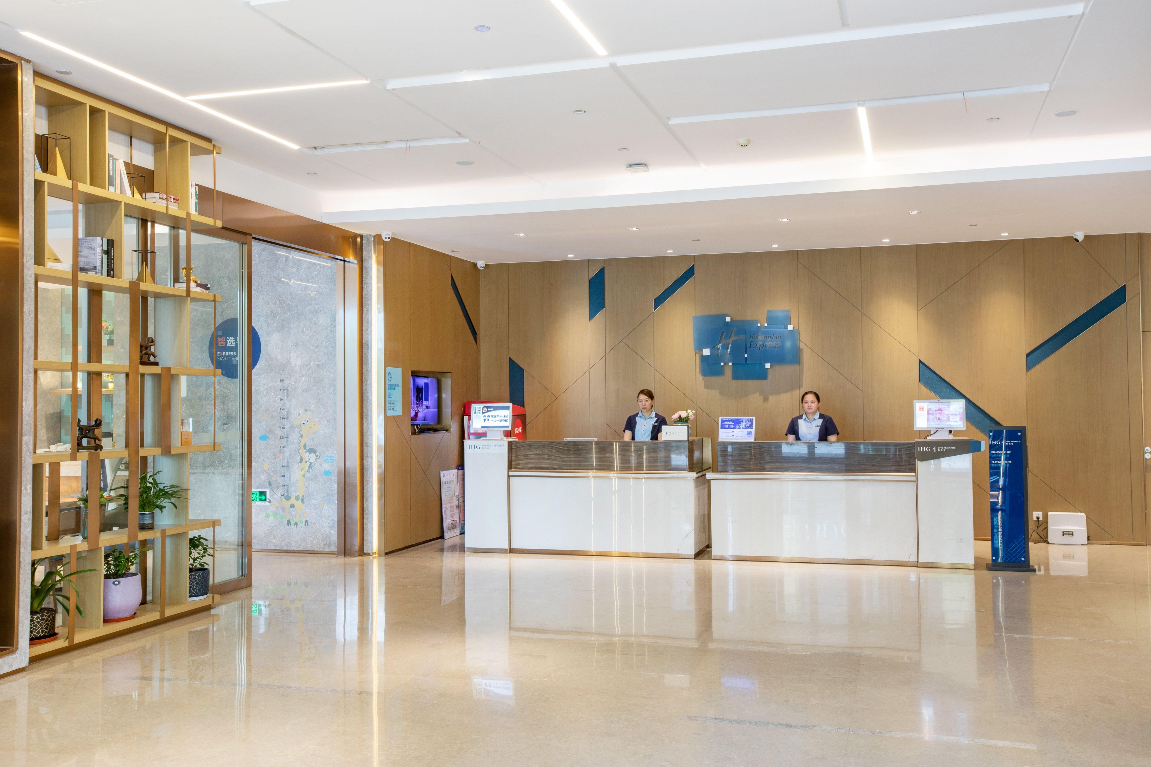 Una moderna zona de recepción con un separador de vidrio, estanterías de madera con plantas y un panel informativo digital.