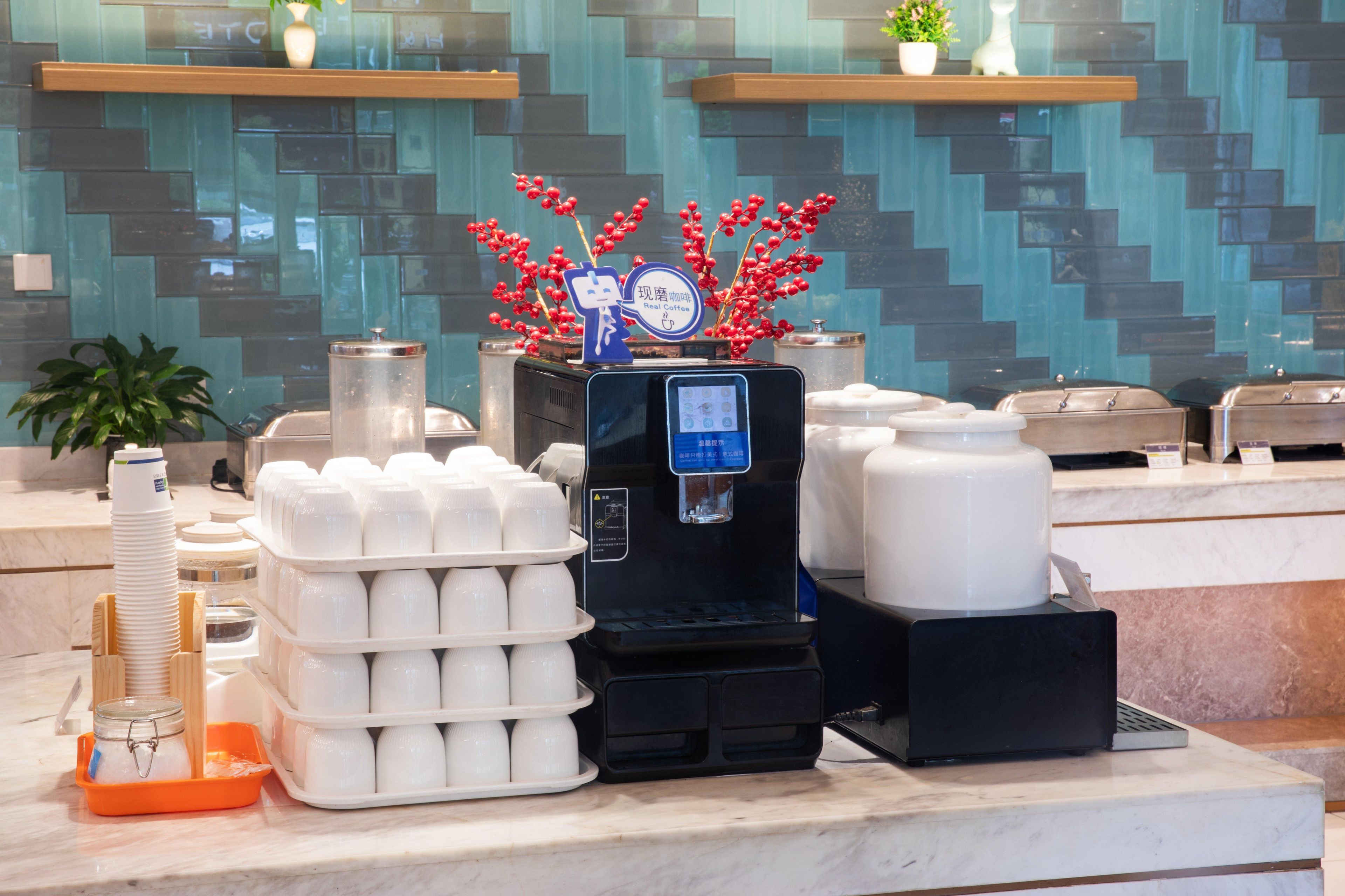 A coffee station with a water dispenser, stacks of disposable cups, and a decorative plant.