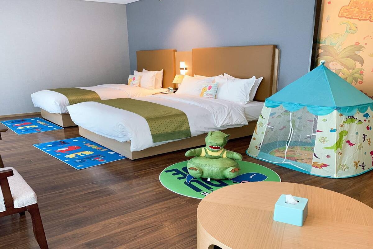 A hotel room with two beds, a play tent, a toy dinosaur, and a small table with a tissue box.
