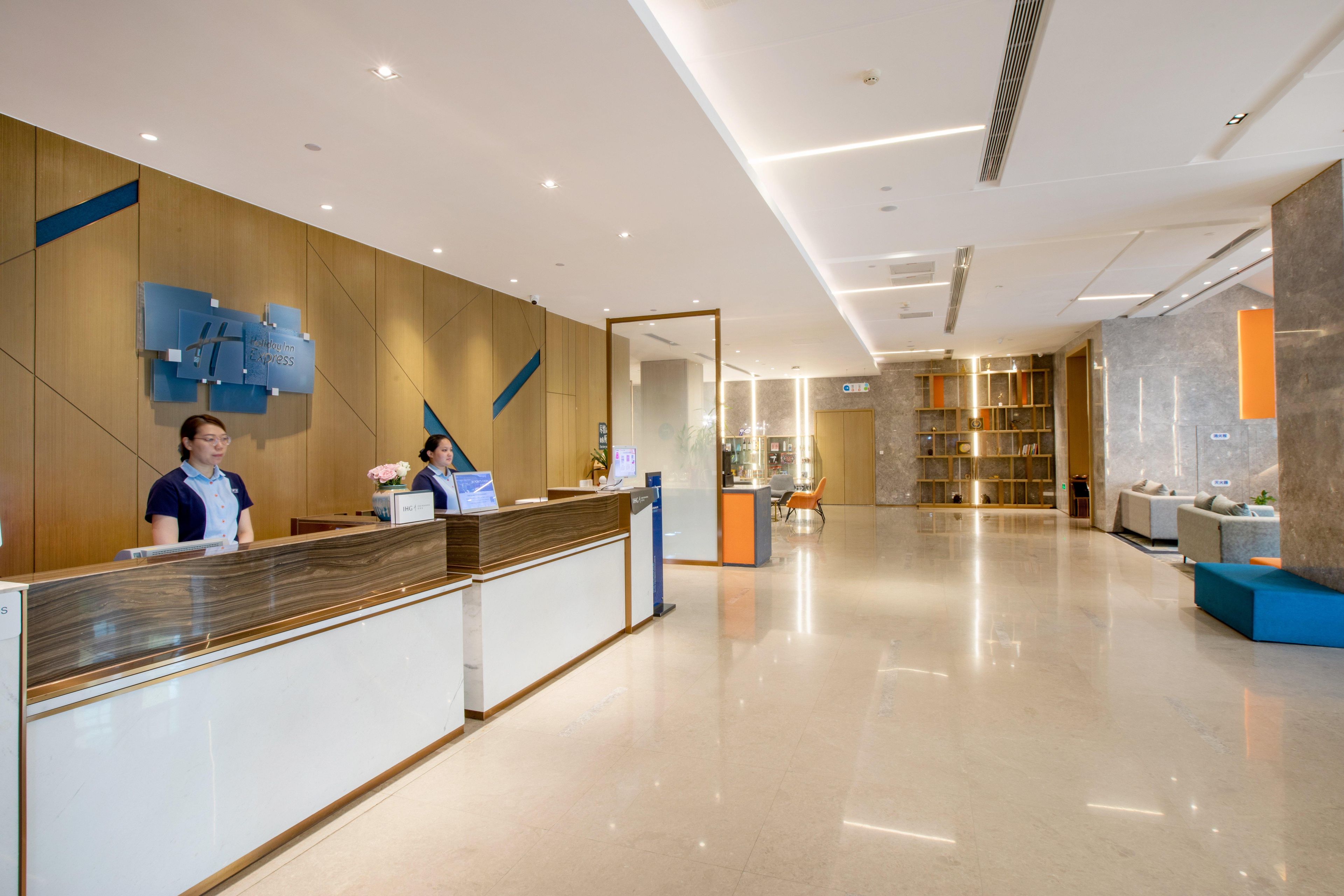 A modern hotel reception area with a wooden reception desk, a digital display, and a seating area with a sofa and chairs.