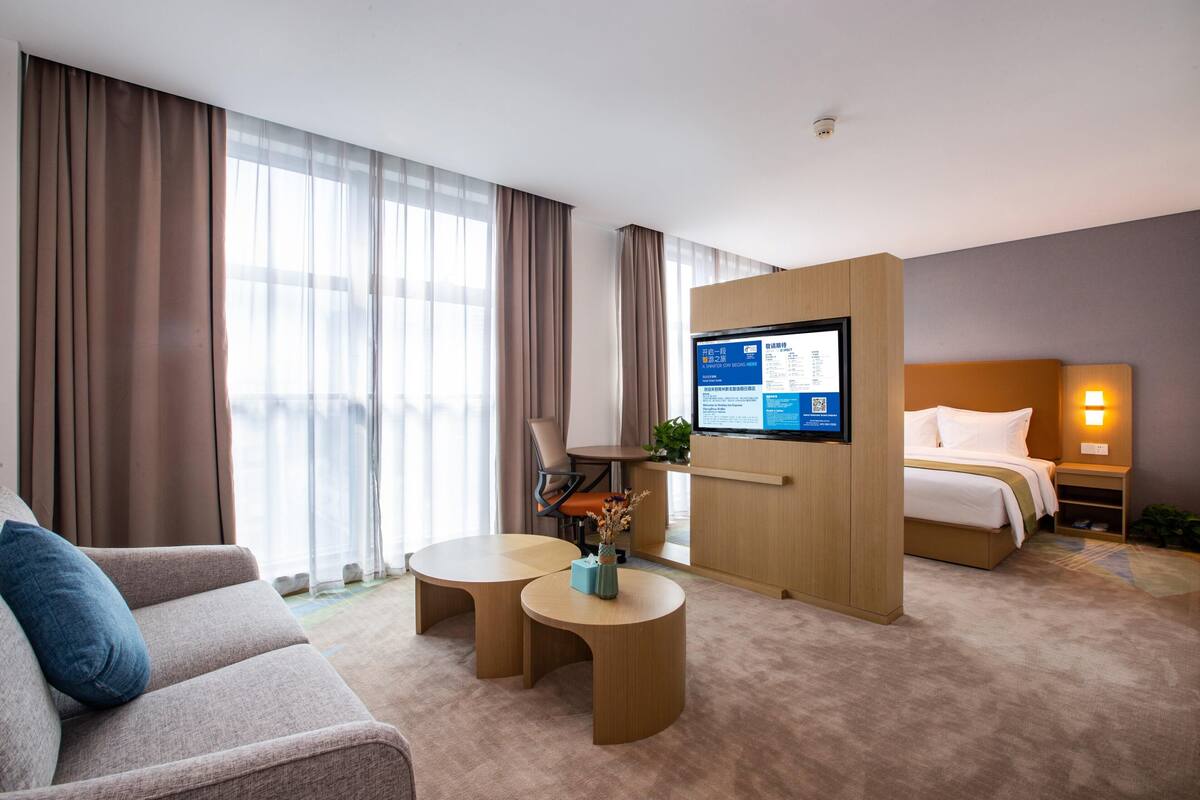 A hotel room with a sofa, two round tables, a bed, a TV mounted on a wooden panel, and a nightstand with a lamp.