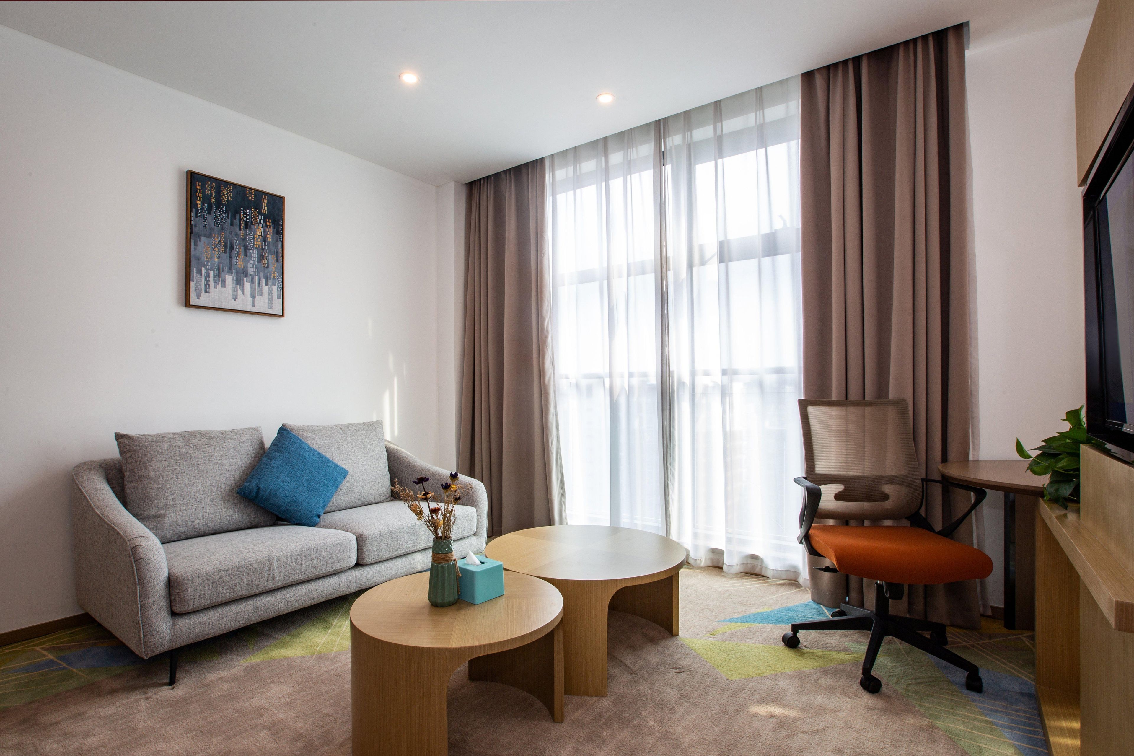 Una habitación de hotel moderna con un sofá gris, dos mesas redondas de madera, una silla de oficina marrón, un televisor sobre un mueble de madera y un amplio ventanal con cortinas traslúcidas.