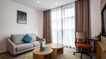 Una habitación de hotel moderna con un sofá gris, dos mesas redondas de madera, una silla de oficina marrón, un televisor sobre un mueble de madera y un amplio ventanal con cortinas traslúcidas.