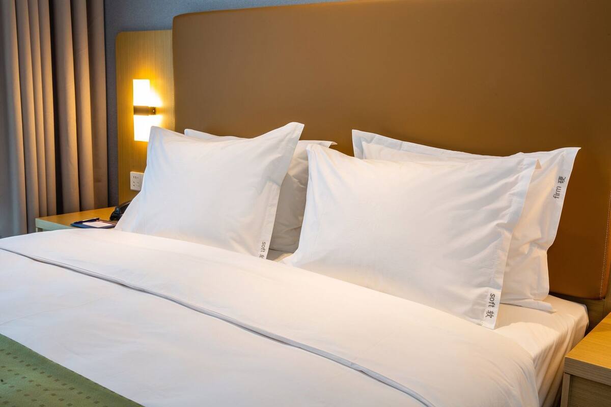 A hotel room with a bed, pillows, a nightstand, a lamp, and a curtain.