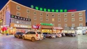 A hotel with multiple floors, a parking lot with several cars, and various storefronts with illuminated signs. - Greentree Inn Liaocheng Gaotang Tianqi Temple Business Center Express Hotel (Liaocheng)