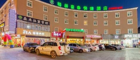 A hotel with multiple floors, a parking lot with several cars, and various storefronts with illuminated signs.