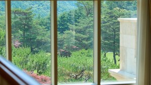A view through a window with a view of a forested area and a building with a stone facade. - Crystal Valley CC - Golden Village (Gapyeong)