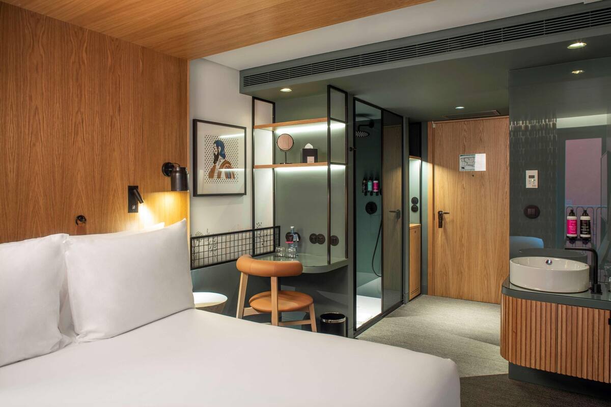 A modern hotel room with a bed, a wooden wall, a glass-enclosed shower, a vanity with a sink, and a small stool.