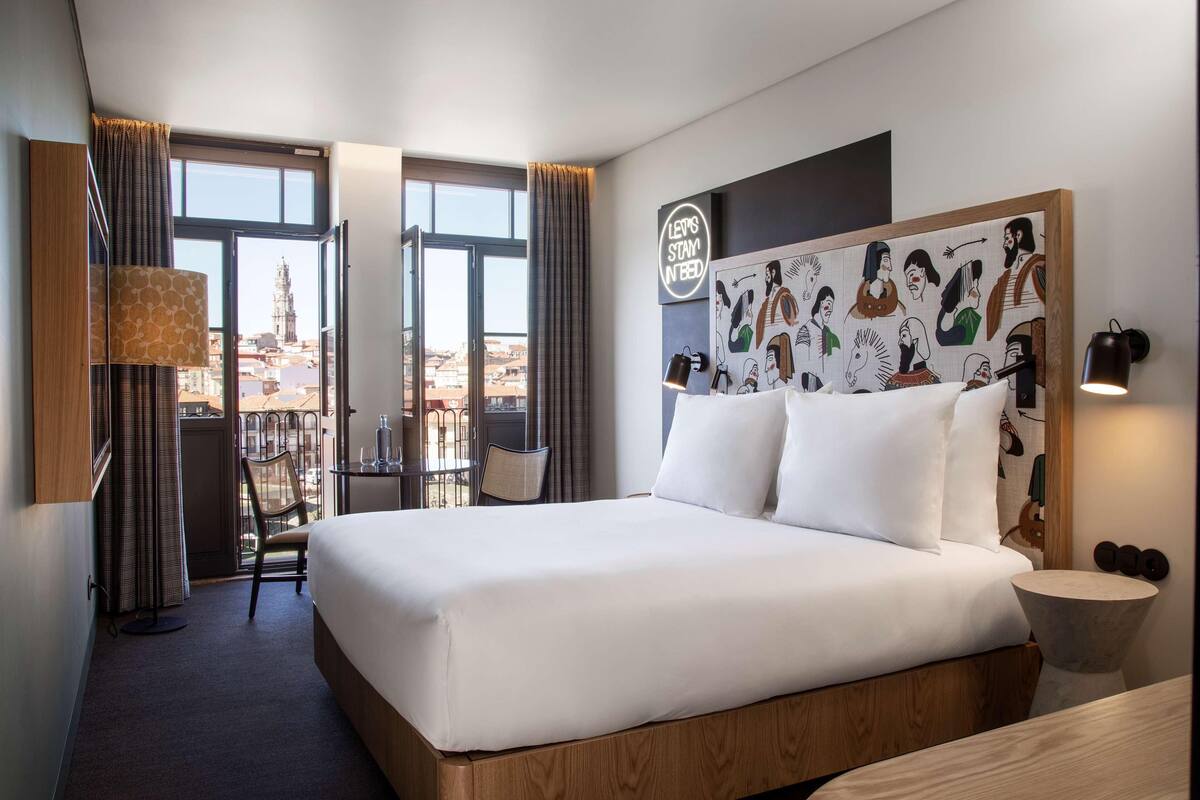 A hotel room with a large bed, a desk, and a chair. There is a view of the city through the windows.