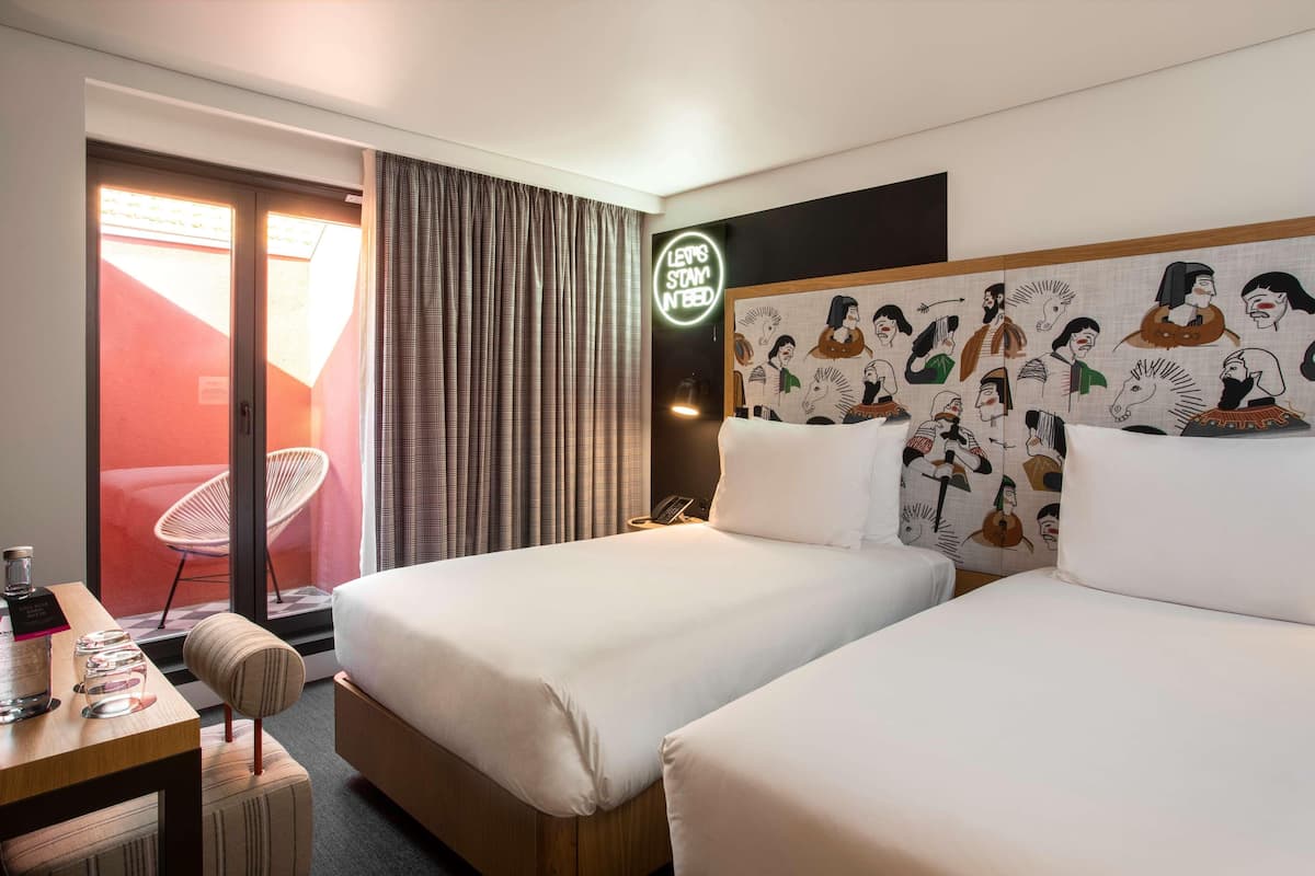 A hotel room with two beds, a desk with a chair, a neon sign, and a wall with a mural.