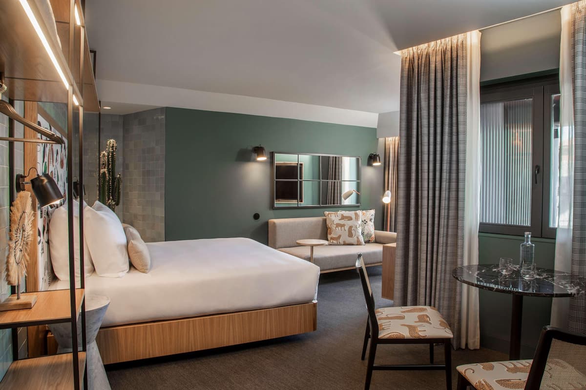 A modern hotel room with a bed, a sofa, a dining table, and a mirror.