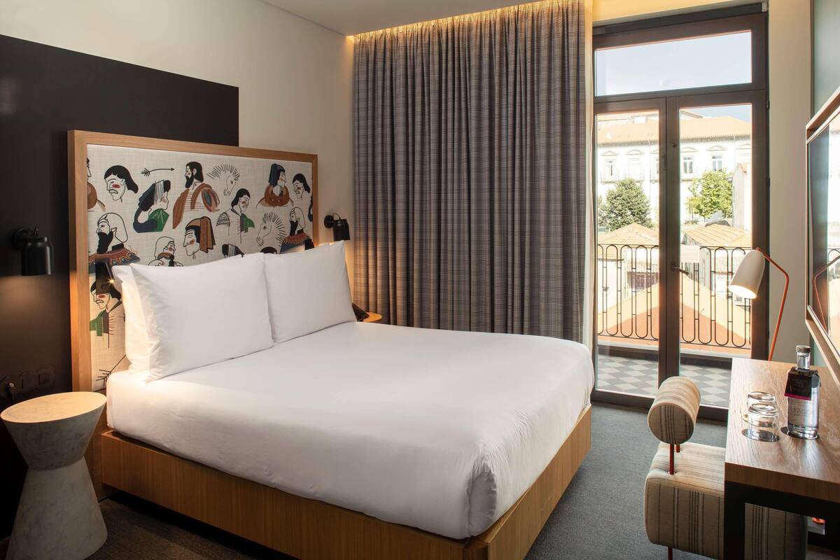 A hotel room with a large bed, a bedside table with a lamp, a small stool, and a view of the outside balcony.