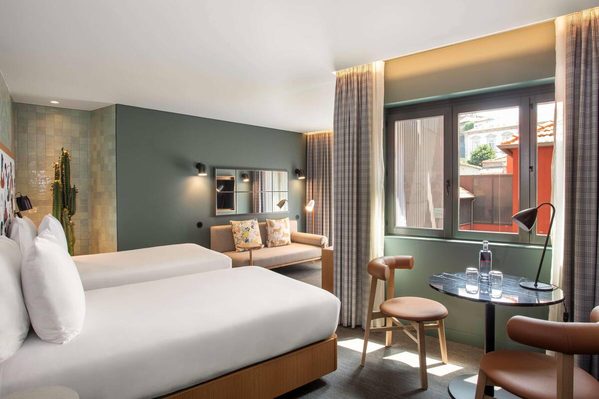 A modern hotel room with a large bed, a sofa, a small table with glasses and a bottle, and a view of the outside.