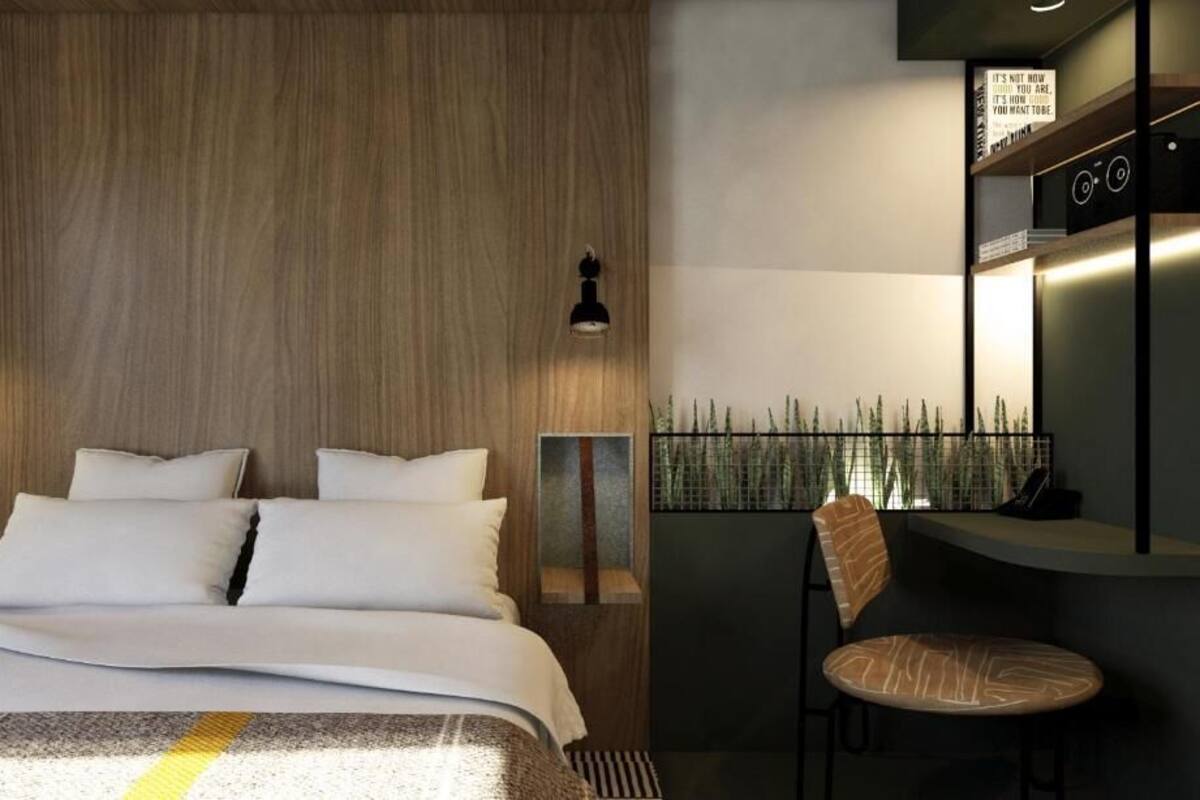 A modern hotel room with a wooden headboard, a bed with white linens, two bedside lamps, a small table with a chair, and a view of a bamboo fence.