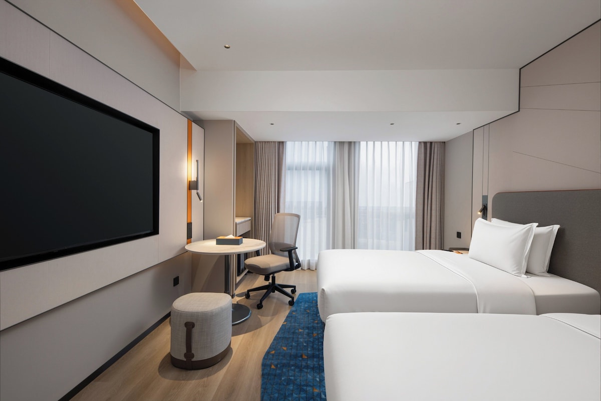 A hotel room with a large flat-screen TV, two beds, a desk, a chair, and a small round ottoman.