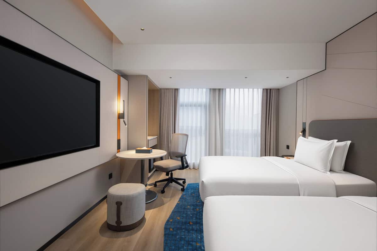 Een hotelkamer met een grote flatscreen televisie, twee bedden, een bureau, een stoel en een klein rond pouf.