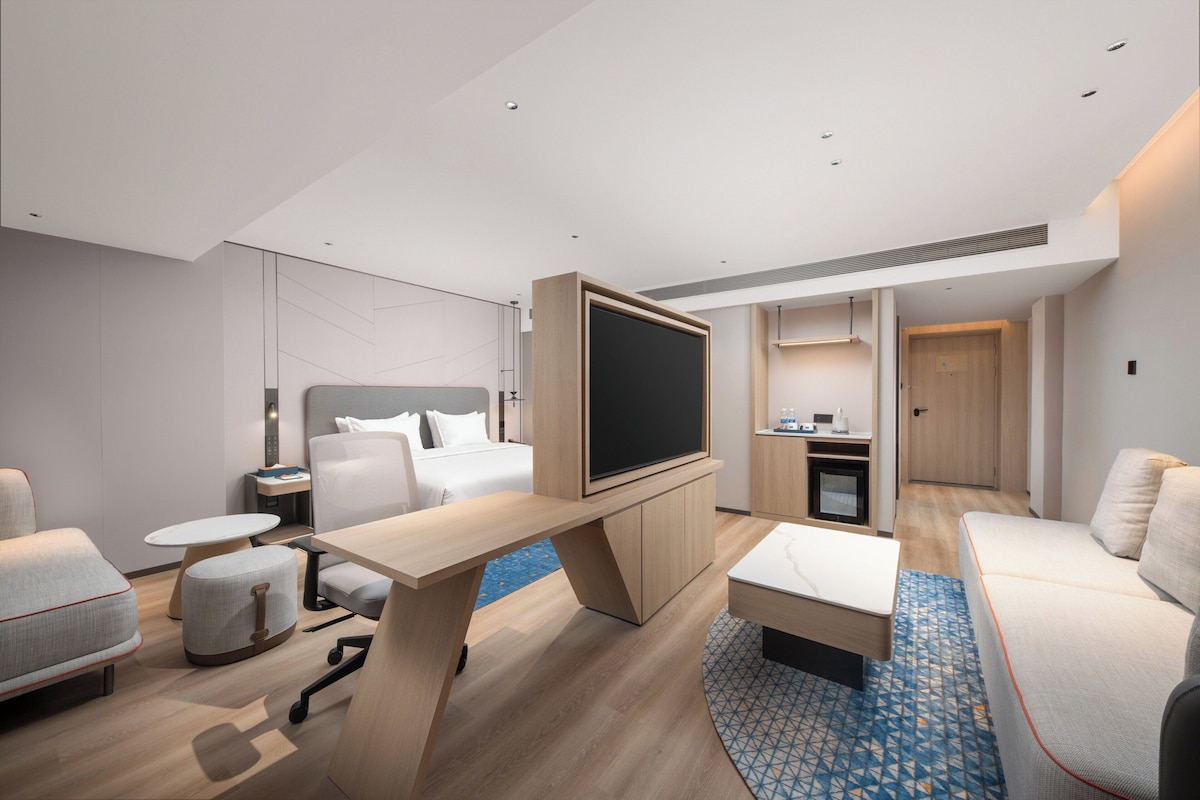 A modern hotel room with a bed, a TV, a desk, a sofa, a small table, and a bathroom visible through an open door.