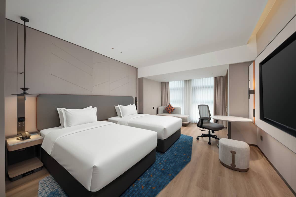 Een hotelkamer met twee bedden, een bureau, een stoel, een televisie en een groot raam met gordijnen.