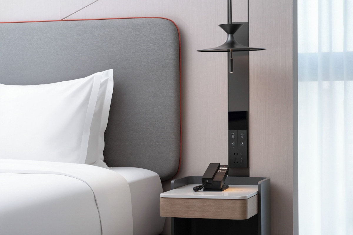 A modern hotel room with a neatly made bed, a bedside table with a phone, and a wall-mounted control panel.