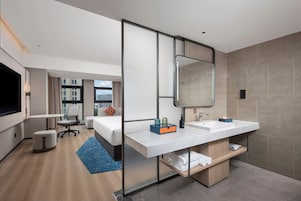 Habitación de hotel moderna con una cama grande, un escritorio, una silla, un baño con espejo y vistas a la ciudad.