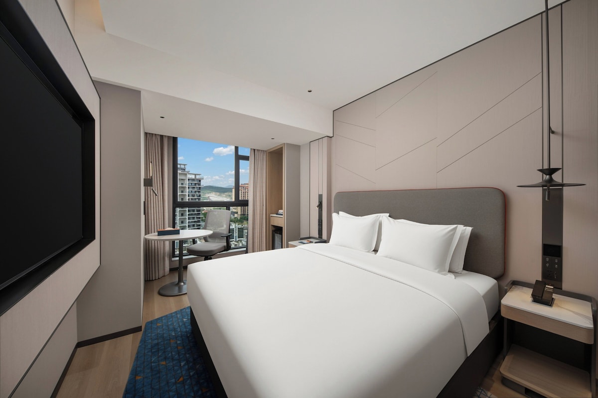 A modern hotel room with a large bed, a desk, a chair, and a TV.