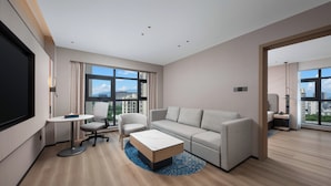 Ein modernes Hotelzimmer mit einer Couch, einem kleinen runden Tisch, einem Sessel, einem Fernseher und großen Fenstern mit Blick auf die Stadt.