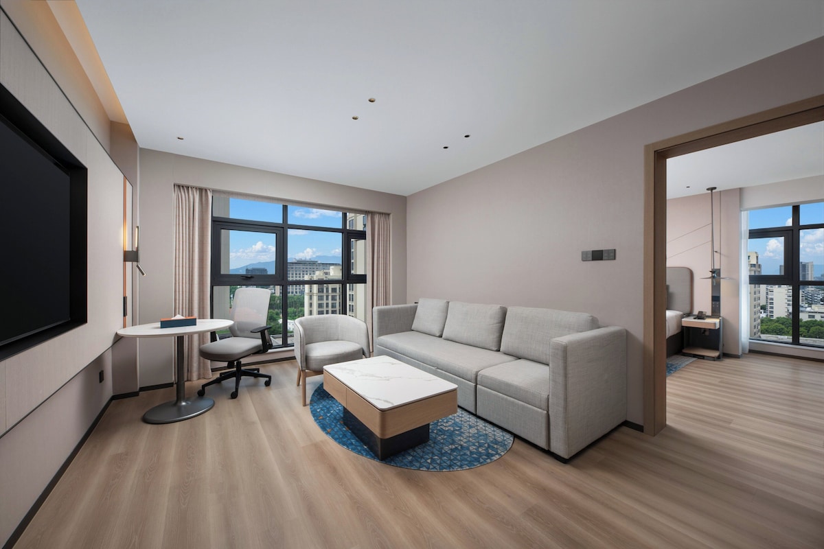 A modern hotel room with a sofa, a small round table, a chair, a TV, and large windows offering a city view.
