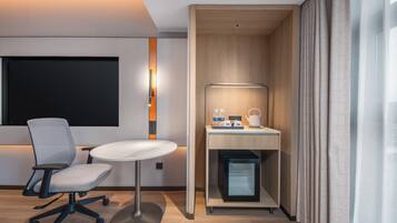 Quarto de hotel moderno com escrivaninha, cadeira, mini-geladeira embutida e televisão na parede.