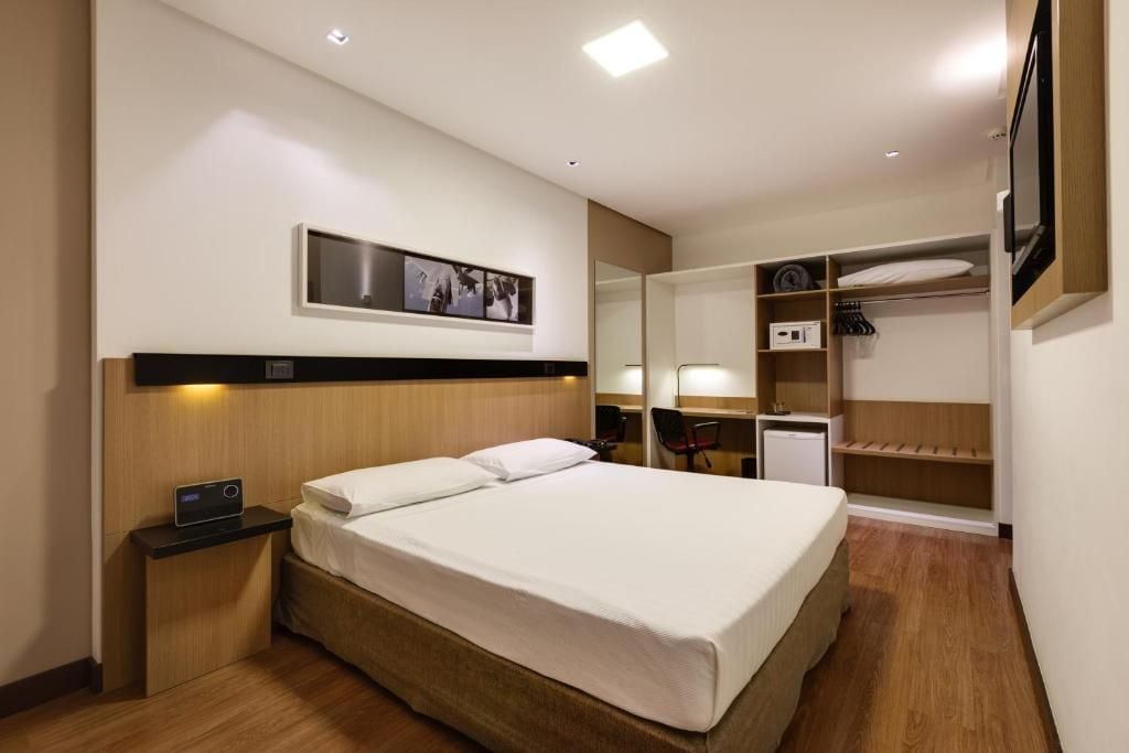Ein Hotelzimmer mit Bett, Nachttisch, Nachttischlampe, Kleiderschrank und einem an der Wand montierten Fernseher.
