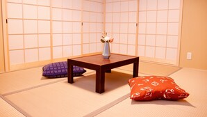 Tatami-mattoilla päällystetty huone, jossa on matala puupöytä, maljakko kukkineen ja kaksi koristetyynyä.