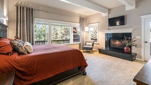 A bedroom with a large bed, a fireplace, a TV mounted on the wall, and a view of the outdoors through large windows. - Royal Oak Manor Lrg private home and yard sleeps 18! (Bend)