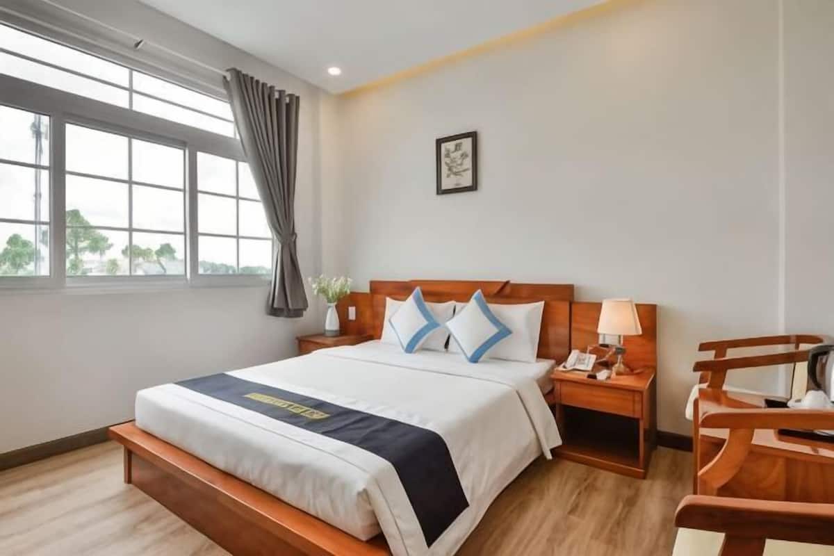 Une chambre d’hôtel avec un lit, une table de chevet, une lampe, une chaise et une fenêtre avec des rideaux.