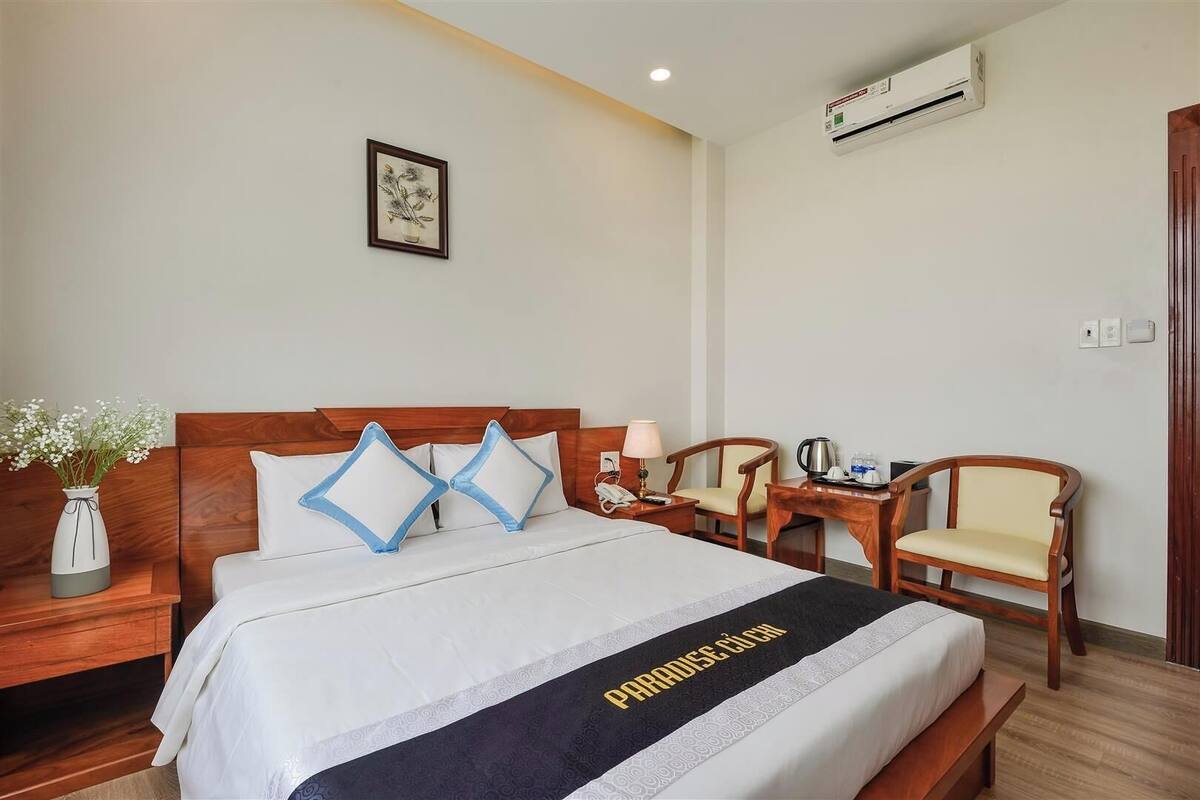 Habitación de hotel con cama, mesita de noche, sillas, escritorio, jarra con flores y equipo de aire acondicionado.