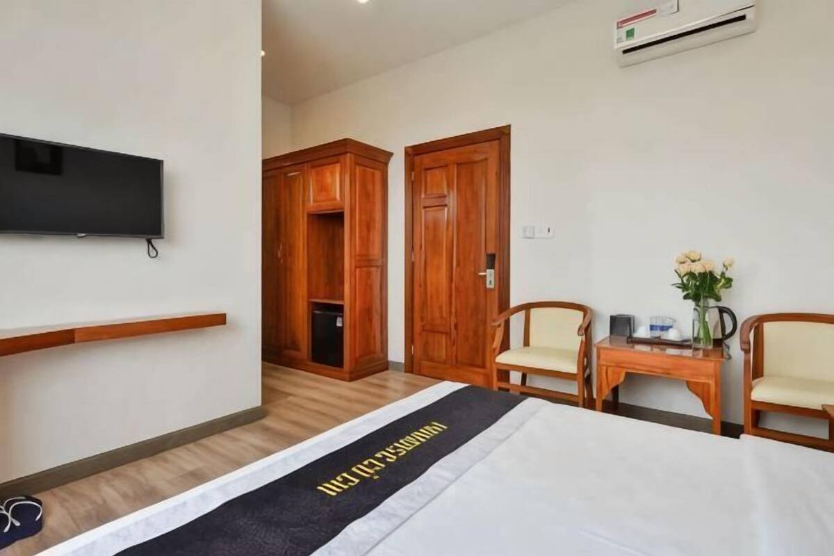 Habitación de hotel con cama, televisor montado en la pared, armarios de madera, una pequeña mesa con sillas y un jarrón con flores.