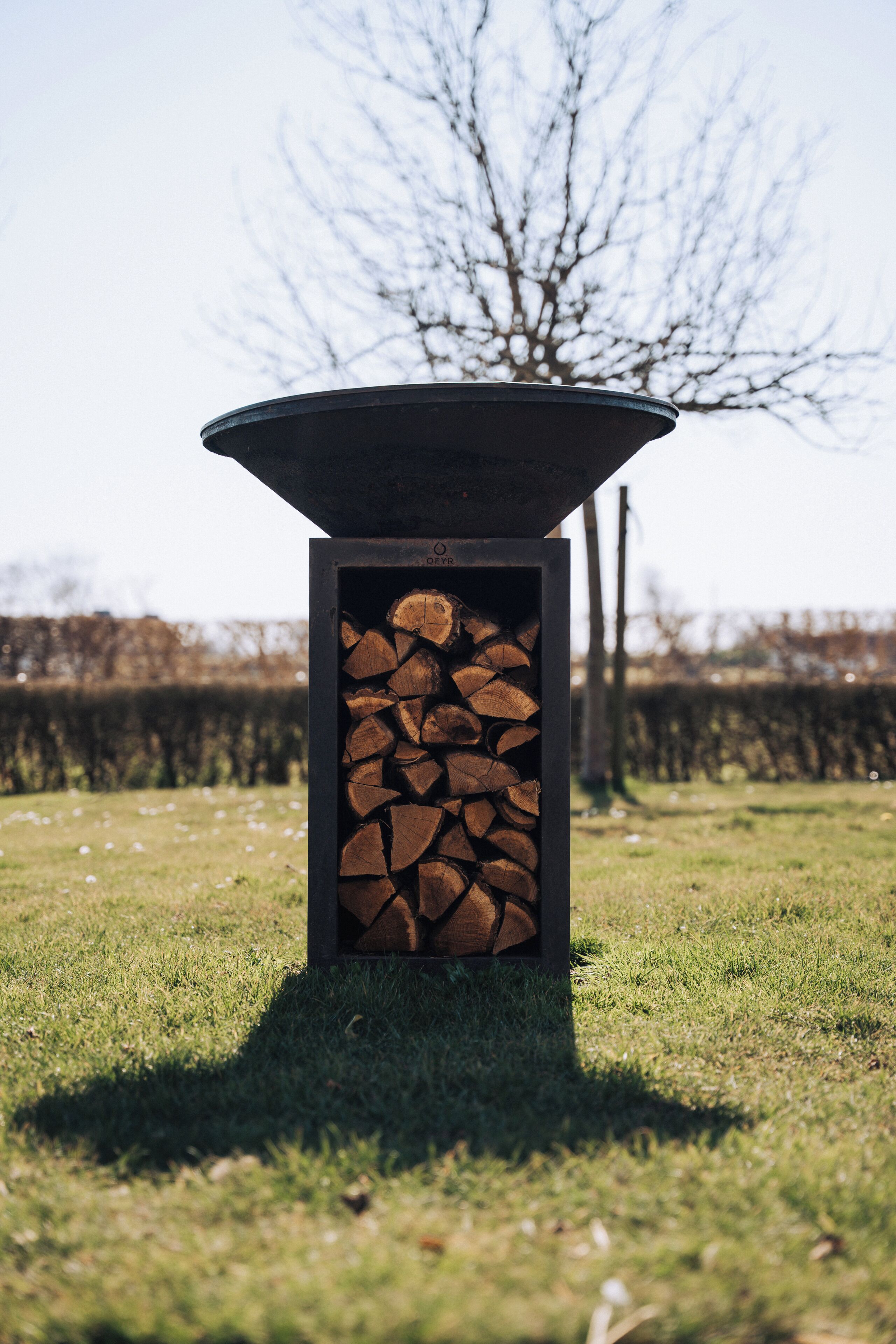 A fire pit with a stack of logs inside, set on a grassy area with a hedge and leafless trees in the background.